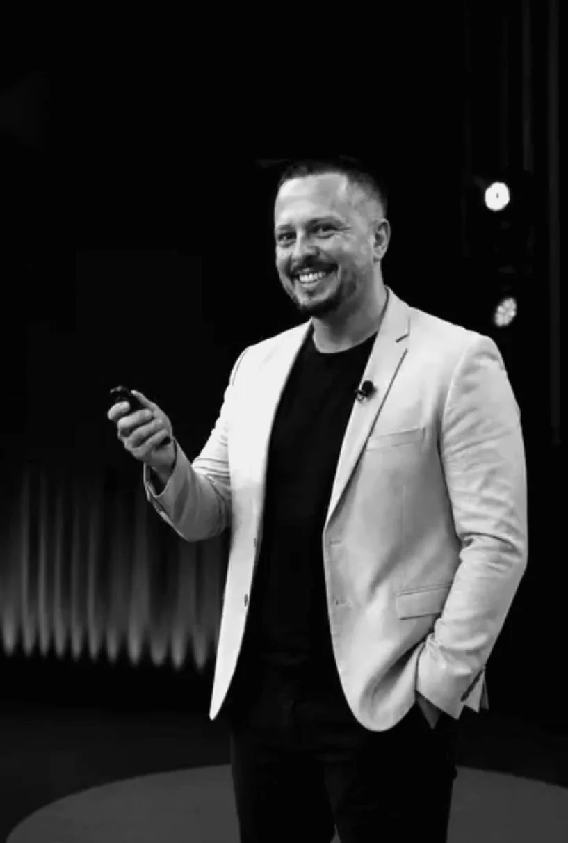O professor sorrindo no palco, em preto e branco, de blazer claro e camiseta escura.