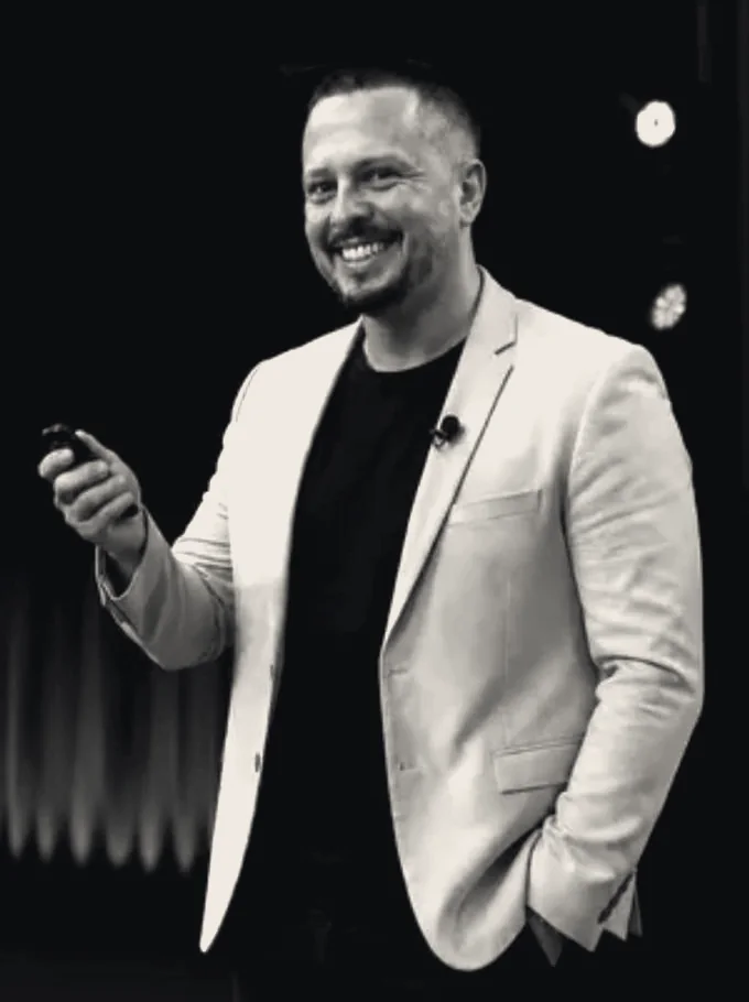 Retrato em preto e branco do professor no palco, sorrindo, de blazer claro.